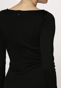 Černý top s dlouhým rukávem a širokým výstřihem, přiléhavý střih a detail na zip na zadní straně, vyrobený z měkkého a elastického materiálu.