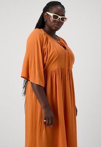 Vestido laranja, de comprimento médio, com tecido texturizado, mangas largas e decote em V. Os acessórios incluem óculos de sol estilo cat-eye de cor clara.