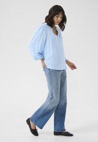 Blouse bleu clair texturé avec un décolleté en V et des manches ballon, associée à un jean droit délavé allant du clair au foncé et des chaussures plates noires.