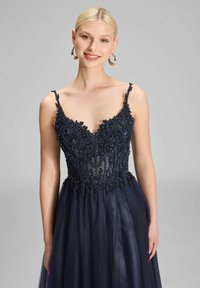 Granatowa sukienka wieczorowa z koronkowym topem, przezroczystym materiałem i delikatnymi ramiączkami. Pełna tiulowa spódnica dodaje objętości i faktury.