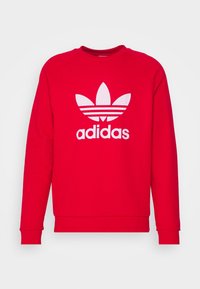 Röd sweatshirt med långa ärmar, rund halsringning och vit Adidas-logga med en trebladig design och horisontella randar under loggan.