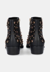 Botines de cuero negro con tachuelas en tono dorado, cierre con cremallera y un tacón bajo cuadrado, con una superficie texturizada y detalles decorativos.