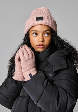 Rosa gestrickte Mütze mit Rippstruktur und Leder-Patch, passend zu handschuhen mit Rippstruktur. Getragen mit einer schwarzen Daunenjacke.