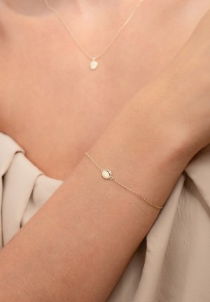 Goldarmband mit einem runden Anhänger, mit einer zarten Kette und glatter Oberfläche, die auf einem hellen Arm auf weichem Stoff ruht.