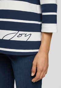 Gestreiftes Shirt in Marineblau und Weiß mit dem Wort "Freude" nahe dem Saum, getragen mit dunkelblauen Jeans, zeigt einen entspannten Arm und eine Hand an der Seite.