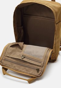 Sac à dos marron ouvert montrant une housse intérieure rembourrée pour ordinateur portable ainsi que des poches en tissu et à filet zippées à l'intérieur du rabat.