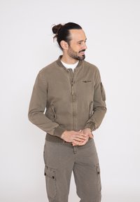 Chaqueta bomber color caqui de un tejido suave y texturizado, con cierre de cremallera, puños acanalados y bolsillos laterales. El modelo lleva pantalones cargo a juego.