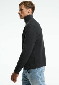 Dunkelgrauer gerippter Pullover mit hohem Kragen, der aus strukturiertem Stoff besteht und eine taillierte Passform hat, kombiniert mit hellblauen Jeans.