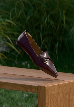 Mocassin en cuir marron avec un détail de nœud flottant au-dessus d’un banc en bois, feuillage vert flou en arrière-plan.