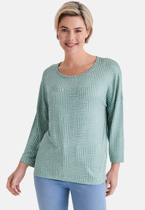 Frau trägt ein hellgrünes Langarmshirt mit weißem, gepunktetem geometrischem Muster und hellblauen Jeans, lächelnd vor weißem Hintergrund.