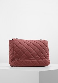 Borsa in tessuto rosa polveroso trapuntata con cuciture a diamante e una tracolla in catena argentata, esposta su un piedistallo bianco su uno sfondo neutro.