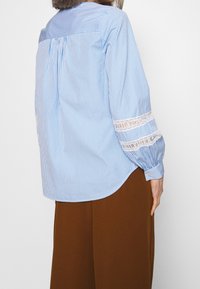 Mujer vistiendo una camiseta de rayas azul y blanca de manga larga con detalles de encaje en las mangas y pantalones de pierna ancha marrones, mostrada desde atrás.