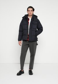 Geacă matlasată bleumarin cu glugă, prevăzută cu închidere cu fermoar, două buzunare frontale și design matlasat; asortată cu pantaloni gri și pantofi negri.