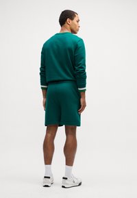 Grøn sweatshirt og matchende shorts, begge med en glat overflade. Sweatshirten har stribede manchetter, og modellen har hvide sneakers på.