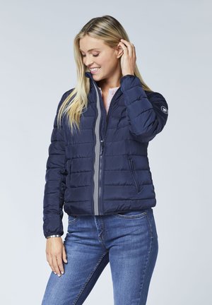 Dunkelblaue Steppjacke mit horizontaler Steppung, durchgehendem Reißverschluss und gerippten Bündchen, kombiniert mit einem hellen Oberteil und blauen Jeans.
