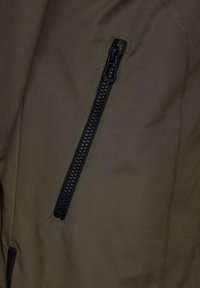 Oliivinvihreä kangas, jossa on sileä pinta, musta vetoketjullinen tasku ja kontrastivärinen ommel. Lähikuvassa vaate.