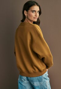Sweatshirt oversize marrone con polsini e orlo a coste, abbinato a jeans patchwork blu chiaro. Lo sfondo presenta un muro tenue.
