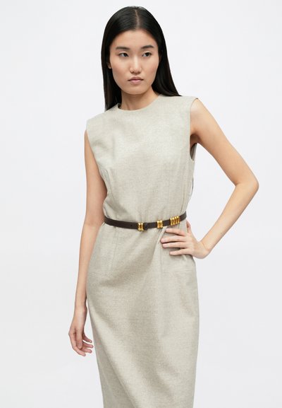 Μπεζ αμάνικο φόρεμα με υφή υφάσματος, εφαρμοστό σχήμα και μπροστινές τσέπες, συνδυασμένο με καφέ ζώνη που έχει χρυσές λεπτομέρειες.