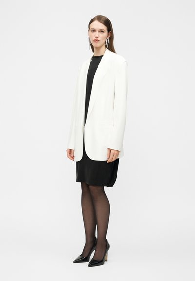 Γυναίκα που στέκεται φορώντας ένα λευκό σακάκι πάνω από ένα μαύρο φόρεμα, διάφανα μαύρα καλσόν, μαύρες ψηλοτάκουνες γόβες και μακριά κρεμαστά σκουλαρίκια.