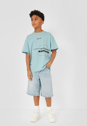 Jongen met kort krullend haar, draagt een lichtblauw oversized voetbalshirt, knielange denim shorts, witte sokken en witte sneakers, staand.
