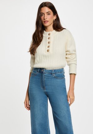 Maglione a coste color crema con colletto alto, caratterizzato da cinque bottoni in tonalità dorata. Abbinato a jeans a gamba larga blu a vita alta.