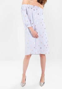 Axel-klänning med blå och vita ränder, prydd med metalliska hjärtmönster och knytärmar. Modellen bär silverfärgade klackar.