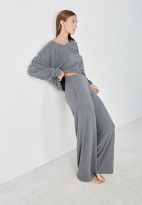 Woman barefoot leaning against white wall, wearing loose grey ribbed knit wide-leg pants and oversized long-sleeve top, looking right.