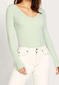 Hellgrünes, geripptes Langarmshirt mit V-Ausschnitt, kombiniert mit hochtaillierten weißen Jeans mit goldfarbenen Beschlägen.