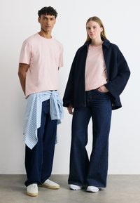 Νεαρός άνδρας και γυναίκα στέκονται όρθιοι, και οι δύο φορούν ροζ μπλούζες με σκούρο μπλε παντελόνια με φαρδιά πόδια· η γυναίκα έχει μια ναυτική μπλε ζακέτα, ο άνδρας ένα ριγέ πουκάμισο δεμένο γύρω από τη μέση.