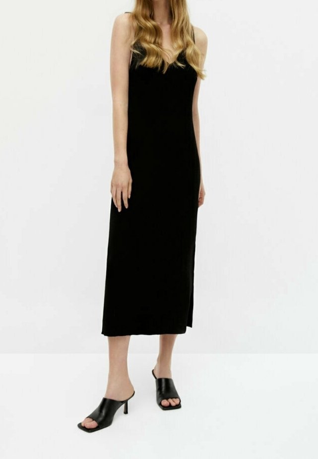 Schwarzes, ärmelloses Kleid mit einer Länge bis zur Mitte der Wade, aus glattem Stoff gefertigt. Kombiniert mit schwarzen, offenen Absatzmules.