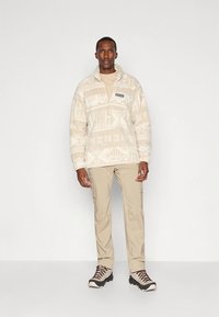 Kreminės spalvos vilnos megztinis su užtrauktuku ant apykaklės, tekstūriniu raštu ir logotipo lipduku. Derinamas su smėlio spalvos krovininėmis kelnėmis ir lengvais žygių batais.