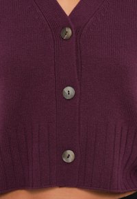 Lila stickad kofta med V-ringning, ribbat nederkant och fyra runda grå knappar fram. Tjock, medelviktig tyg.