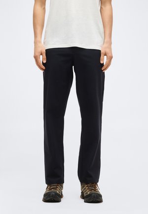 Personne portant un pantalon noir à jambes droites et des chaussures de randonnée marron, debout contre un fond blanc avec les mains détendues sur les côtés.