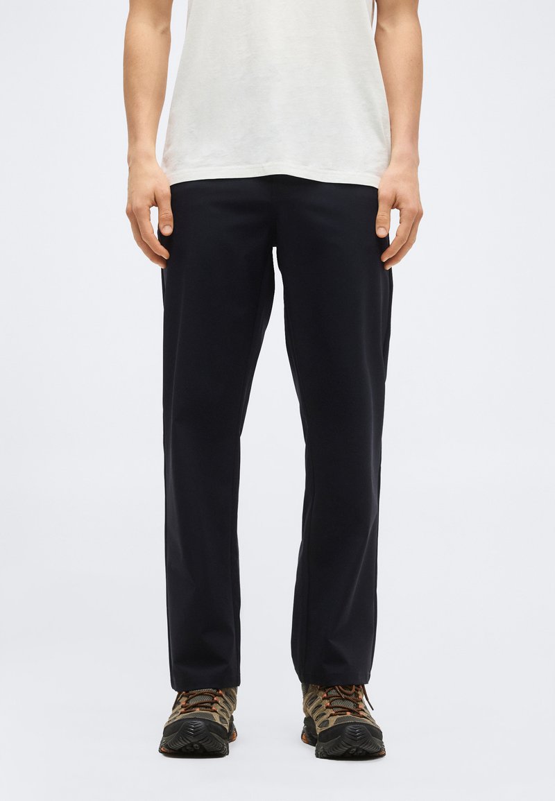 Personne portant un pantalon noir à jambes droites et des chaussures de randonnée marron, debout contre un fond blanc avec les mains détendues sur les côtés.