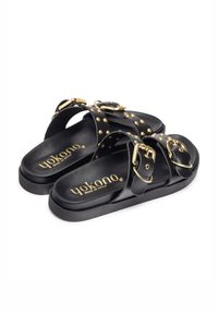 Zwarte leren sandalen met gouden gespen en studs, met een platte zool en brede banden voor ondersteuning. De merknaam is zichtbaar op de voetbed.