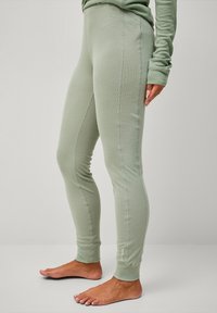 Person, die enganliegende, hellgrüne Leggings und ein passendes langärmliges Oberteil trägt, barfuß vor einem schlichten, hellen Hintergrund steht.