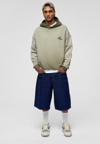 Mann, der einen beigen und grünen Kapuzenpullover mit dem Text "Stay Moment" trägt, navyblaue lange Jeansshorts, weiße Socken, Turnschuhe und eine beigen Kappe, die rückwärts getragen wird.