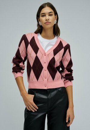 Joven mujer con cárdigan de rombos rosa y burdeos sobre top blanco, pantalones de cuero negro y pendientes plateados, de pie con una mano en el bolsillo.