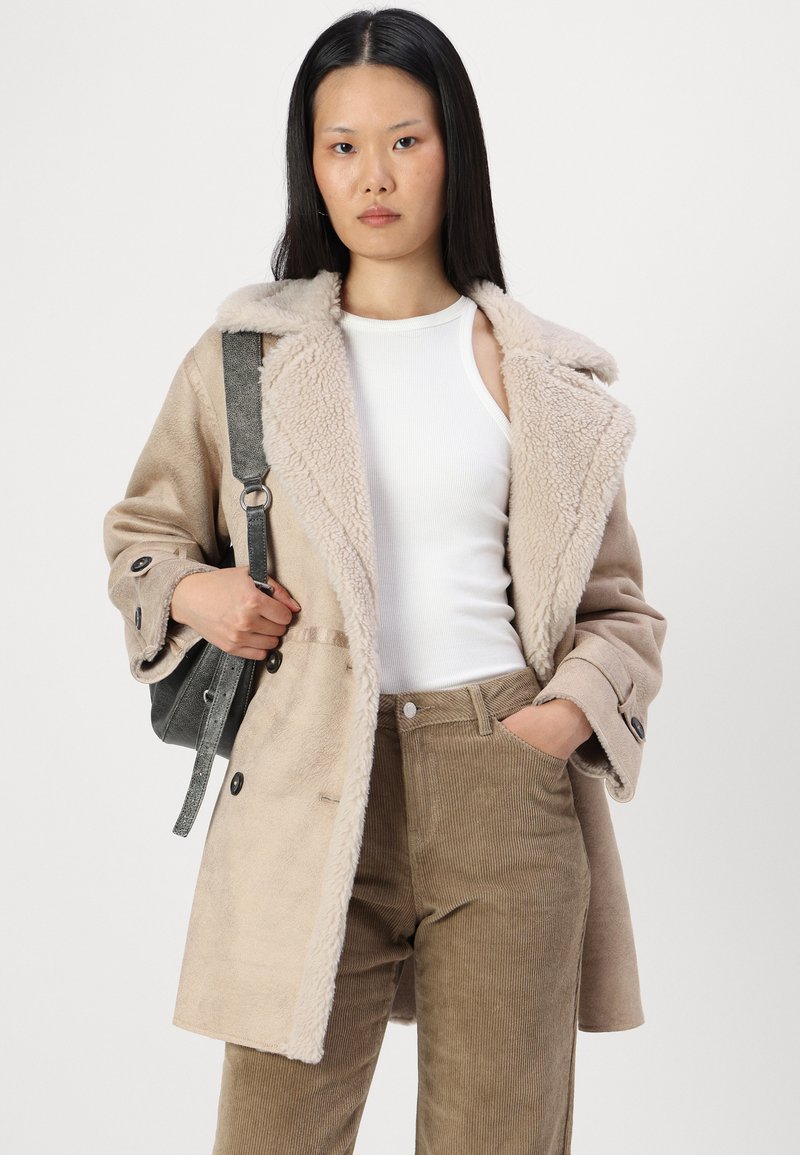 Manteau beige en fausse fourrure avec grands revers, boutons noirs, porté sur un débardeur blanc et un pantalon en velours côtelé marron, tenu avec une sangle de sac grise.