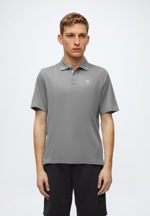Jeune homme portant un polo gris à manches courtes avec des boutons, debout devant un fond clair uni.