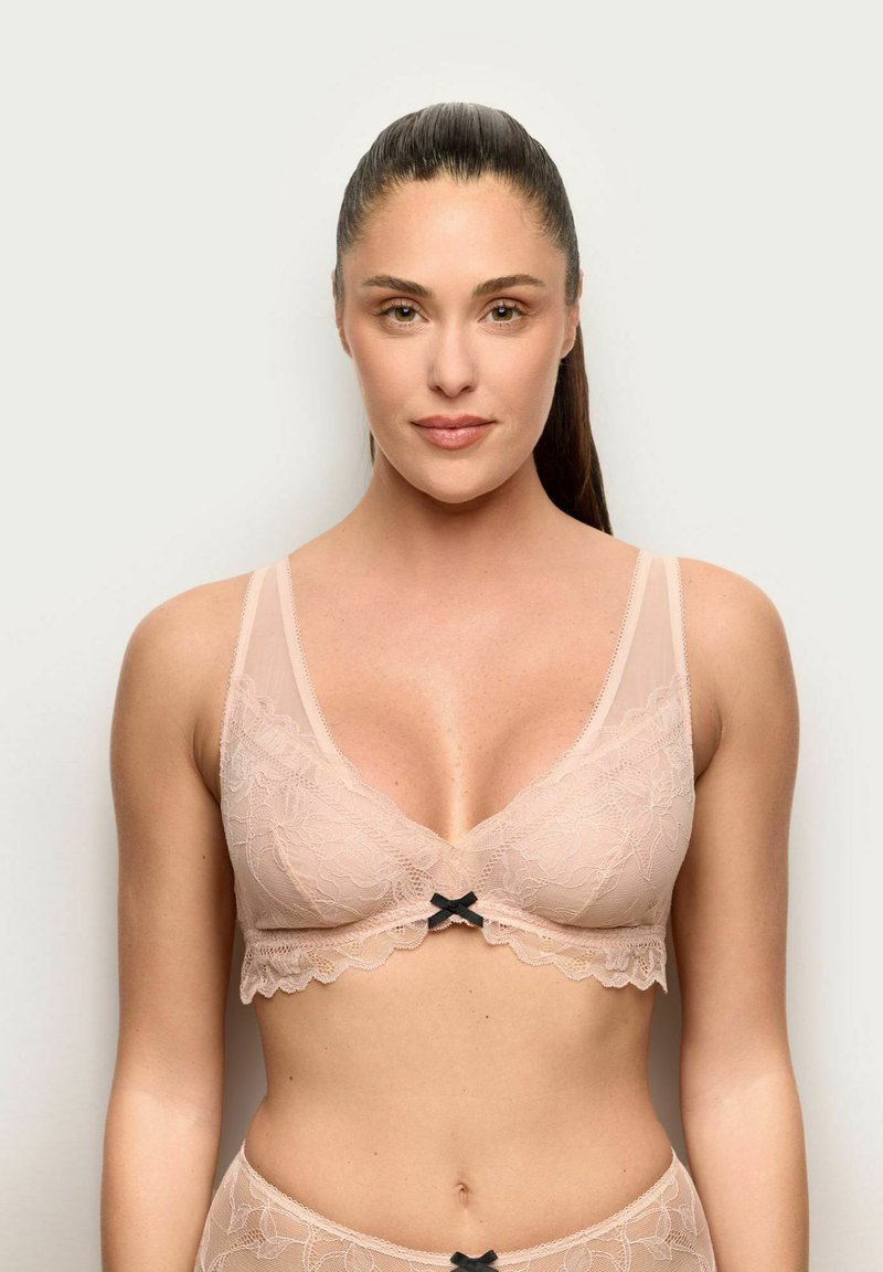 Vrouw met lang donker haar in een naaktkleurige kanten beha en bijpassende slip, staand tegen een effen lichte achtergrond, naar voren gericht.