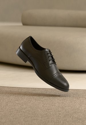 Zapato masculino de vestir de cuero negro con detalles brogue flotando sobre un suelo alfombrado beige y un sofá beige al fondo.