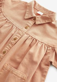 Chemise boutonnée en velours côtelé rose clair avec col, empiècement froncé, poche avant et boutons en bois sur fond blanc.