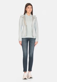 Giacca leggera azzurro chiaro con cappuccio, con zip frontale, abbinata a una polo azzurro chiaro, jeans scuri e sneaker pastello.