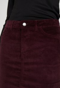 Gros plan sur une personne portant un haut noir et une jupe en velours côtelé violet foncé taille haute avec un bouton en argent et des poches avant.