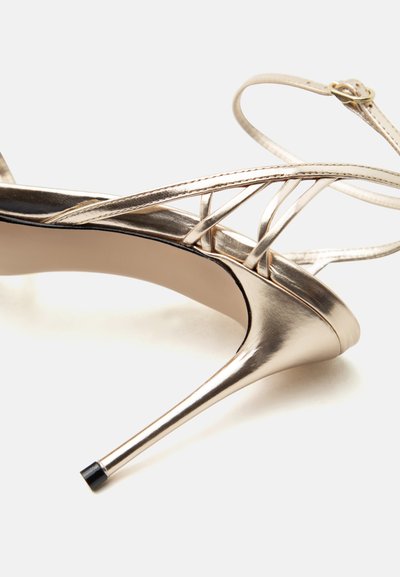 Χρυσό μεταλλικό σανδάλι στιλέτο με λεπτούς λουράκια που διασταυρώνονται και αγκράφα στον αστράγαλο, πάνω σε λευκό φόντο.