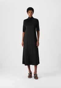 Rochie midi neagră cu guler înalt și mâneci scurte, având o textură netedă. Asortată cu sandale negre cu cataramă. Design simplu și elegant.