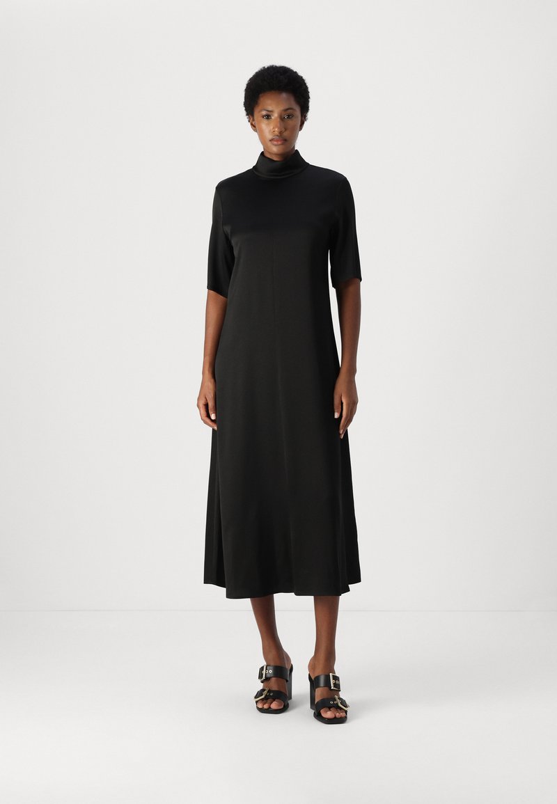 Rochie midi neagră cu guler înalt și mâneci scurte, având o textură netedă. Asortată cu sandale negre cu cataramă. Design simplu și elegant.