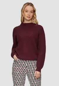 Burgunderfarbener gerippter Pullover mit hohem Kragen und V-förmigem Muster; getragen mit gemusterten schwarzen, weißen und roten Hosen.