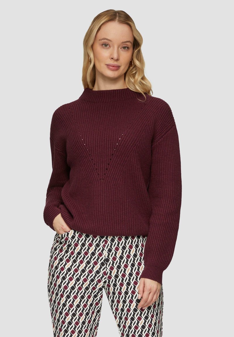Burgunderfarbener gerippter Pullover mit hohem Kragen und V-förmigem Muster; getragen mit gemusterten schwarzen, weißen und roten Hosen.